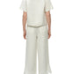 A person stands facing away, wearing the Simkhai Louisa Camp Shirt and wide-leg white pants with embroidered hems, paired with white sandals.
