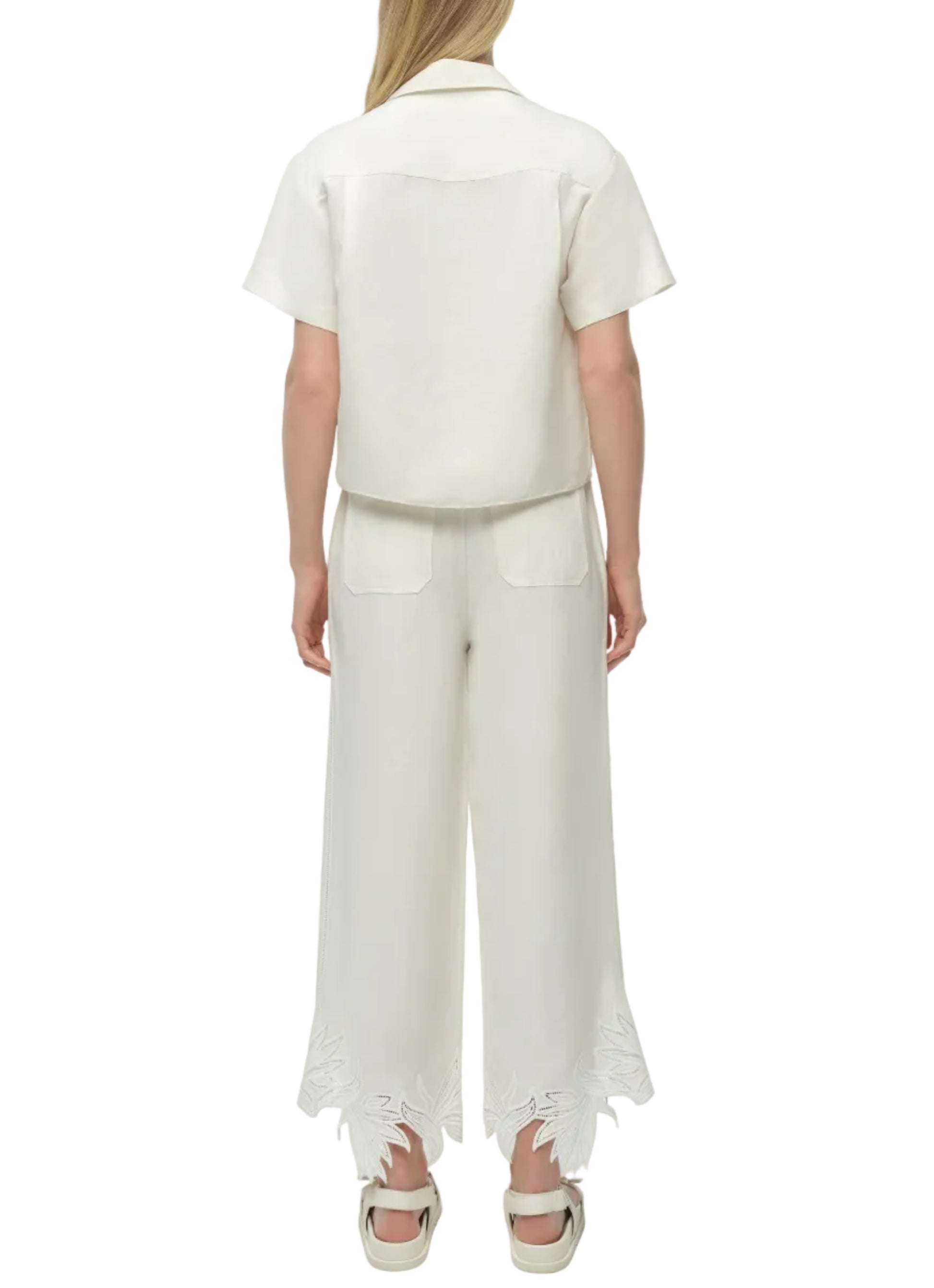 A person stands facing away, wearing the Simkhai Louisa Camp Shirt and wide-leg white pants with embroidered hems, paired with white sandals.