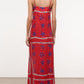 A person stands facing away, wearing the Staud Laura Linen Maxi Dress—an ankle-length red summer dress with blue floral patterns and adjustable spaghetti straps—paired with flat sandals against a plain background.