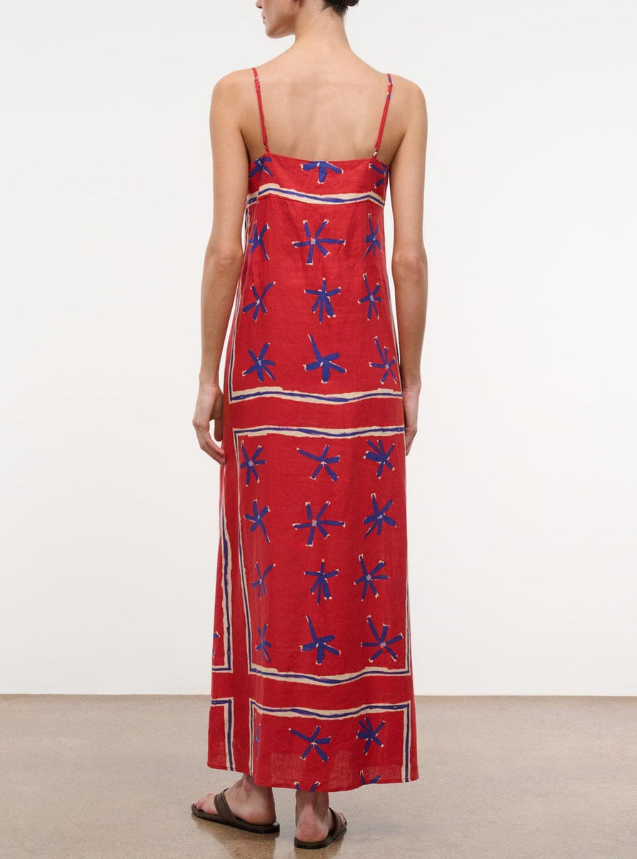 A person stands facing away, wearing the Staud Laura Linen Maxi Dress—an ankle-length red summer dress with blue floral patterns and adjustable spaghetti straps—paired with flat sandals against a plain background.