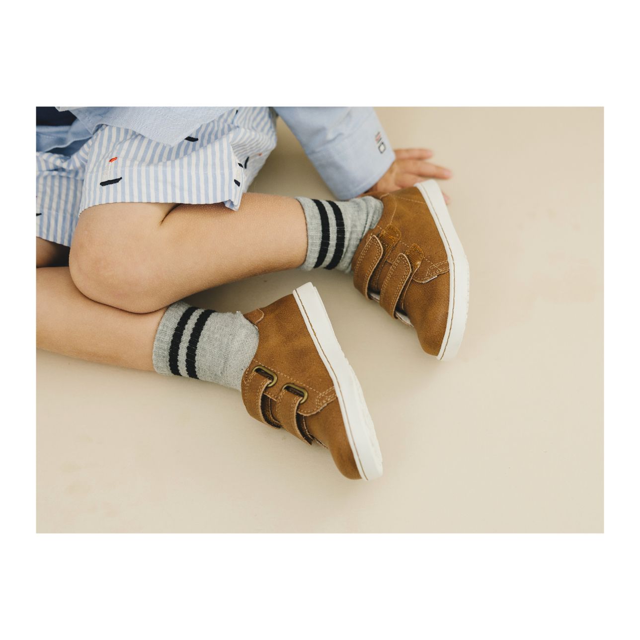 A child in a blue striped shirt sits on the floor, wearing gray socks and brown L'Amour Shoes Boys' Kyle Double Strap Sneakers with a comfy memory foam leather sockbed.