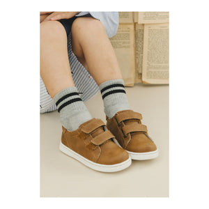 A child in striped grey socks wears L'Amour Shoes' Boys' Kyle Double Strap Sneakers, featuring a comfortable leather sockbed, sitting with knees bent and open books in the background.