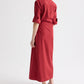 A person faces away, wearing the TWP New Earl Dress—a long red superfine cotton shirt dress with rolled-up sleeves and flat sandals—posed against a plain white background.