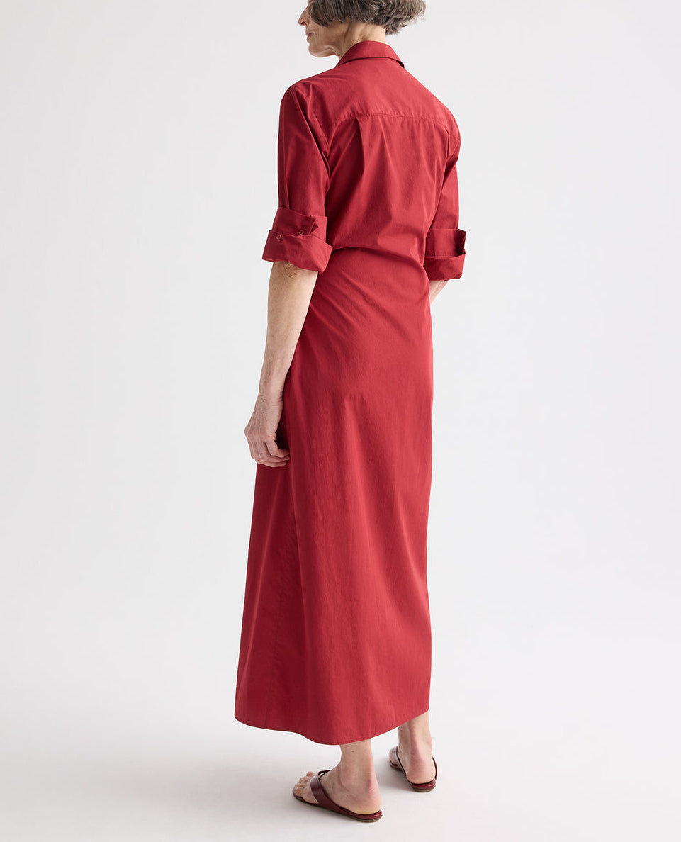 A person faces away, wearing the TWP New Earl Dress—a long red superfine cotton shirt dress with rolled-up sleeves and flat sandals—posed against a plain white background.