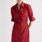 A woman in the TWP New Earl Dress—a red, long-sleeve superfine cotton shirt dress with an asymmetric wrap-front, rolled-up sleeves, and a chest pocket—stands against a plain background.