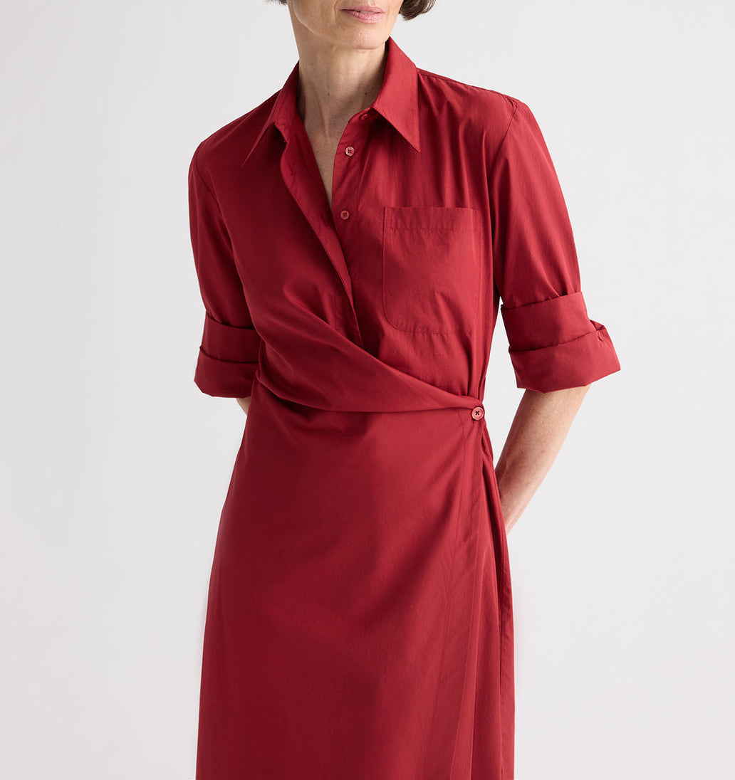 A woman in the TWP New Earl Dress—a red, long-sleeve superfine cotton shirt dress with an asymmetric wrap-front, rolled-up sleeves, and a chest pocket—stands against a plain background.