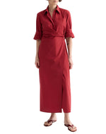 A person wears the TWP New Earl Dress—a long-sleeved, red asymmetric wrap-front shirt dress with a collar and button detail—paired with brown flip-flops, standing against a white background.