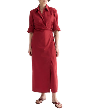 A person wears the TWP New Earl Dress—a long-sleeved, red asymmetric wrap-front shirt dress with a collar and button detail—paired with brown flip-flops, standing against a white background.