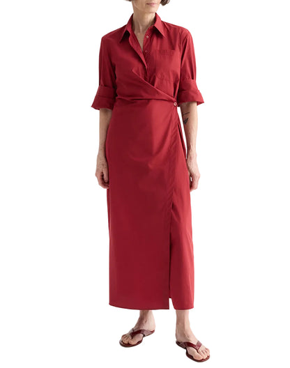 A person wears the TWP New Earl Dress—a long-sleeved, red asymmetric wrap-front shirt dress with a collar and button detail—paired with brown flip-flops, standing against a white background.