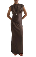 A person wearing the TWP On The Side Dress, a sleeveless, floor-length brown ruched maxi with subtle draping, and a necklace, standing with hands behind their back.