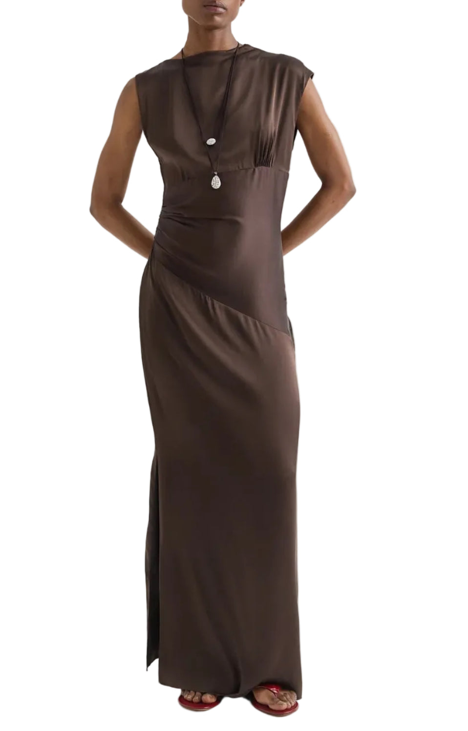 A person wearing the TWP On The Side Dress, a sleeveless, floor-length brown ruched maxi with subtle draping, and a necklace, standing with hands behind their back.
