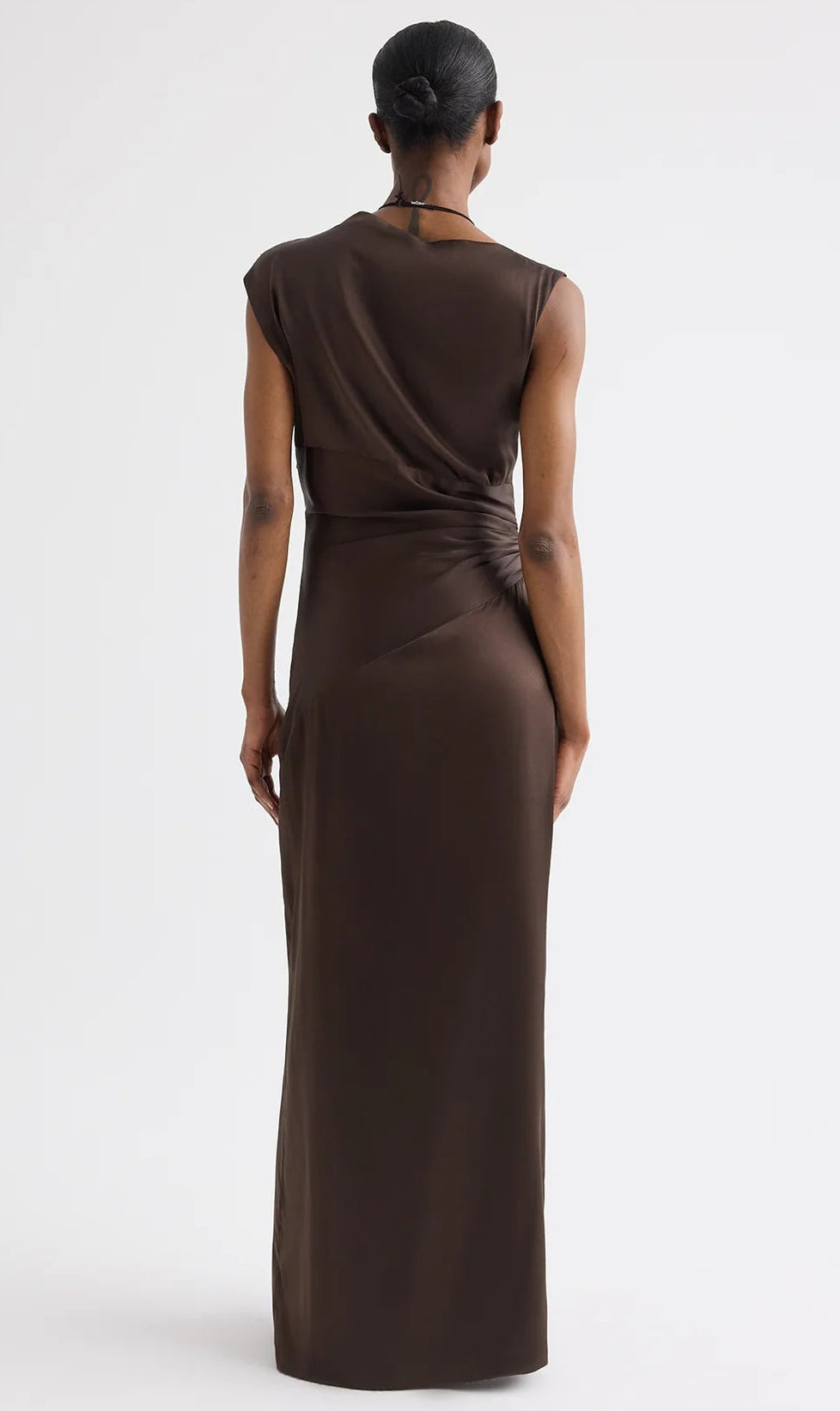 A person faces away, wearing TWP's On The Side Dress—a sleeveless, dark brown silk charmeuse dress with subtle draping and gathered detailing on one side.