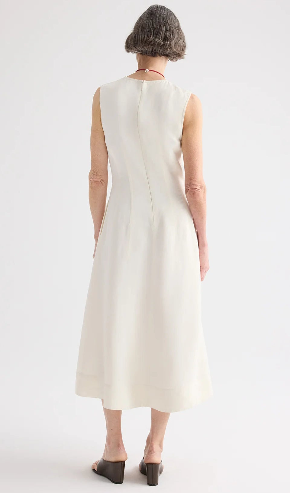 A person with short gray hair faces away, wearing the TWP Crew Neck Coated Viscose Linen Dress, a sleeveless cream A-line style, paired with heeled sandals.