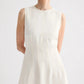 A woman with short gray hair stands with her arms behind her back, wearing the TWP Crew Neck Coated Viscose Linen Dress, featuring a sleeveless design and A-line silhouette, against a plain background.