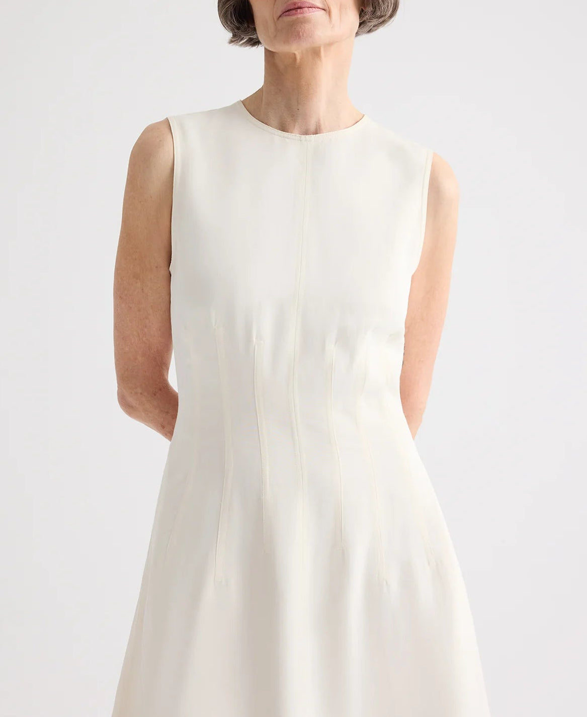 A woman with short gray hair stands with her arms behind her back, wearing the TWP Crew Neck Coated Viscose Linen Dress, featuring a sleeveless design and A-line silhouette, against a plain background.