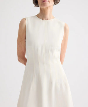 A woman with short gray hair stands with her arms behind her back, wearing the TWP Crew Neck Coated Viscose Linen Dress, featuring a sleeveless design and A-line silhouette, against a plain background.