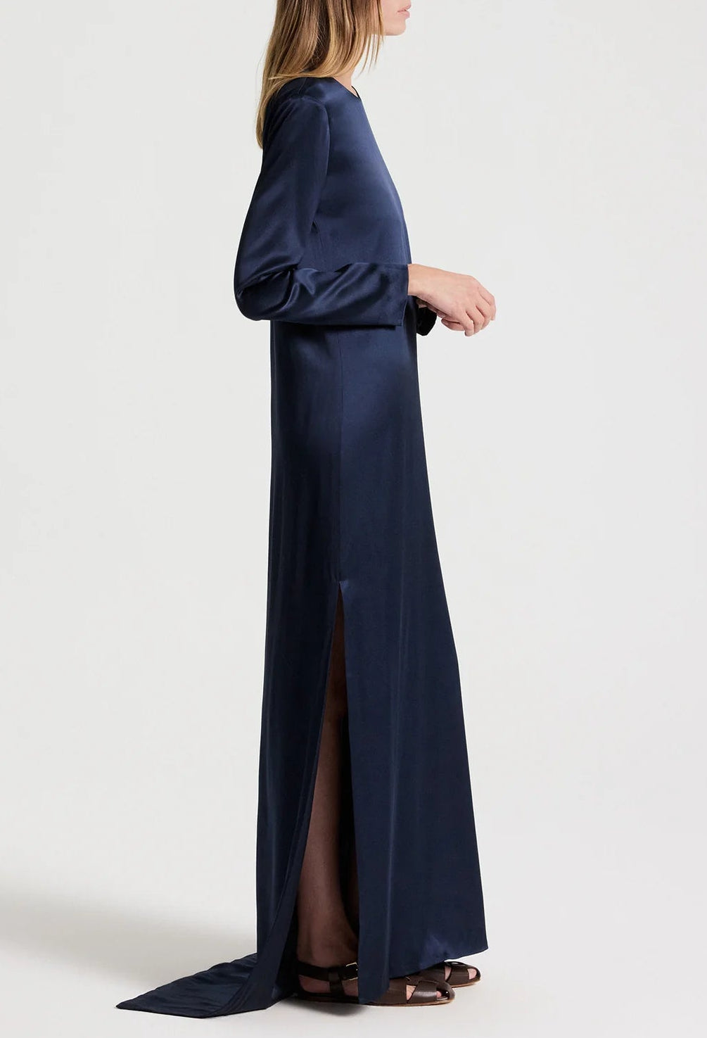 A person wears a long-sleeved, navy blue floor-length TWP Nightfall Dress with a side slit and dark sandals.