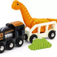 A black toy train from the BRIO World Dinosaur Circle Set, connected to a white train car by a BRIO magnet, is carrying an orange long-necked dinosaur figure. In front of the train, there is a small green bush-like object.