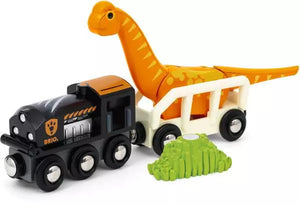 A black toy train from the BRIO World Dinosaur Circle Set, connected to a white train car by a BRIO magnet, is carrying an orange long-necked dinosaur figure. In front of the train, there is a small green bush-like object.