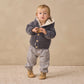A toddler stands on a textured rug wearing checkered Quincy Mae Giovani Pants with an elastic waistband, a gray hooded jacket with an adjustable drawstring, and tan shoes, holding a small object in both hands.