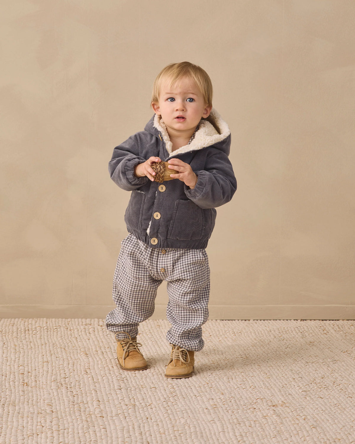 A toddler stands on a textured rug wearing checkered Quincy Mae Giovani Pants with an elastic waistband, a gray hooded jacket with an adjustable drawstring, and tan shoes, holding a small object in both hands.
