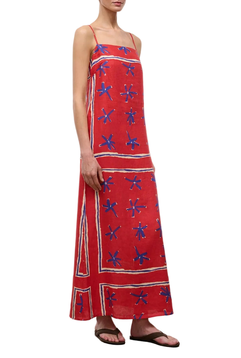 A person wearing the Staud Laura Linen Maxi Dress, a sleeveless red summer dress with blue star-like patterns and adjustable spaghetti straps, paired with black sandals.