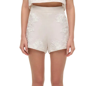 A person wearing Simkhai's Sammie Short—high-waisted white linen shorts with delicate soutache embroidery—and a matching cropped top, arms relaxed at their sides.