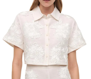 A woman wears the Simkhai Callie Camp Shirt—a short-sleeve, cropped linen top with floral soutache embroidery—paired with matching high-waisted bottoms for an effortless vacation vibe.