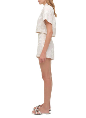 A woman stands in profile wearing the Simkhai Callie Camp Shirt—a light short-sleeve linen shirt—paired with matching shorts featuring delicate soutache embroidery, styled with slip-on sandals against a plain white background.