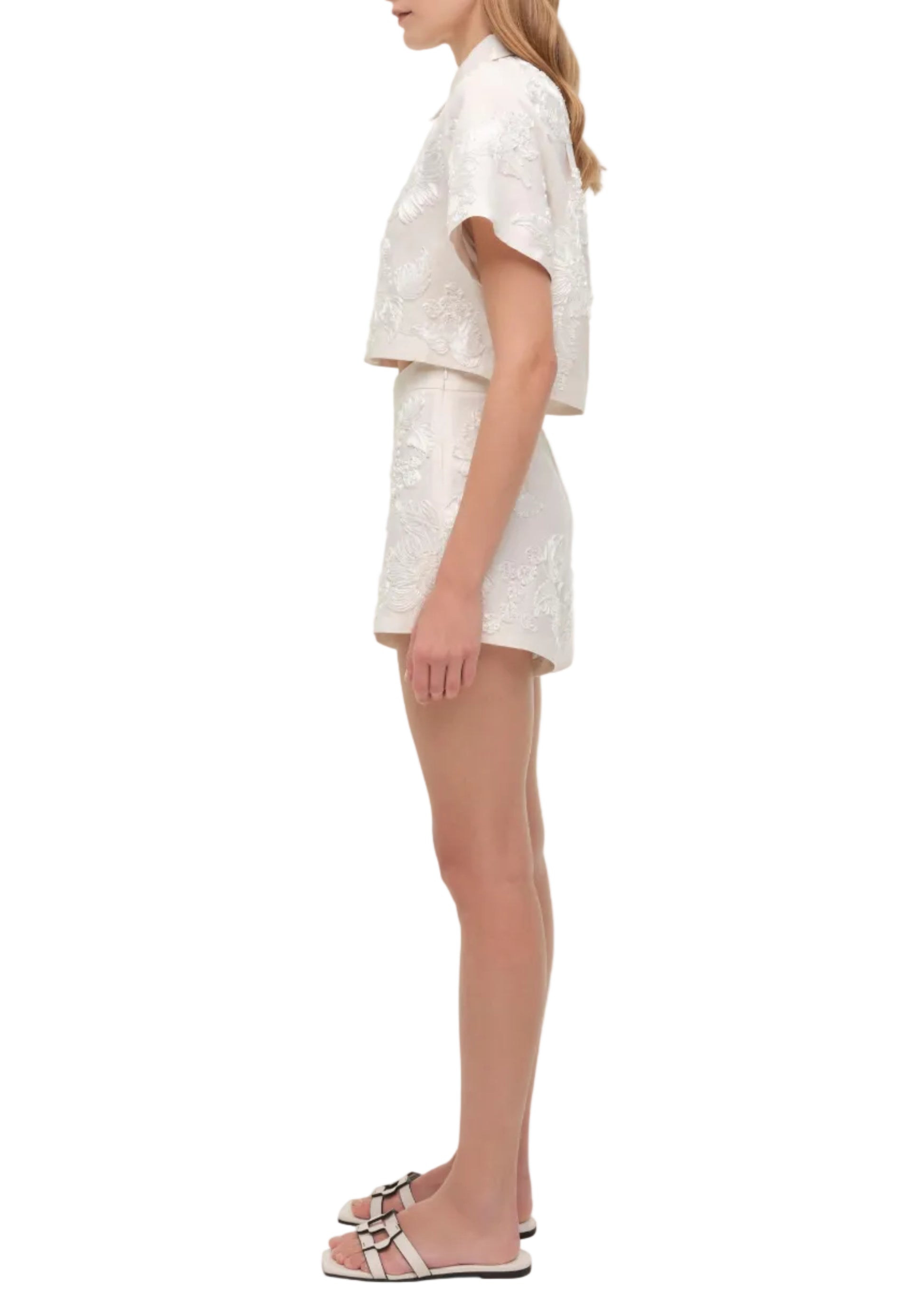 A woman stands in side view wearing Simkhai’s Sammie Short embroidered set with a short-sleeve top, styled with light pink sandals for a breezy look.
