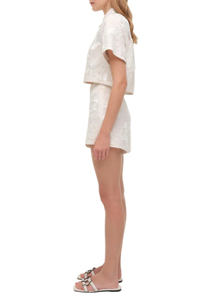 A woman stands in side view wearing Simkhai’s Sammie Short embroidered set with a short-sleeve top, styled with light pink sandals for a breezy look.