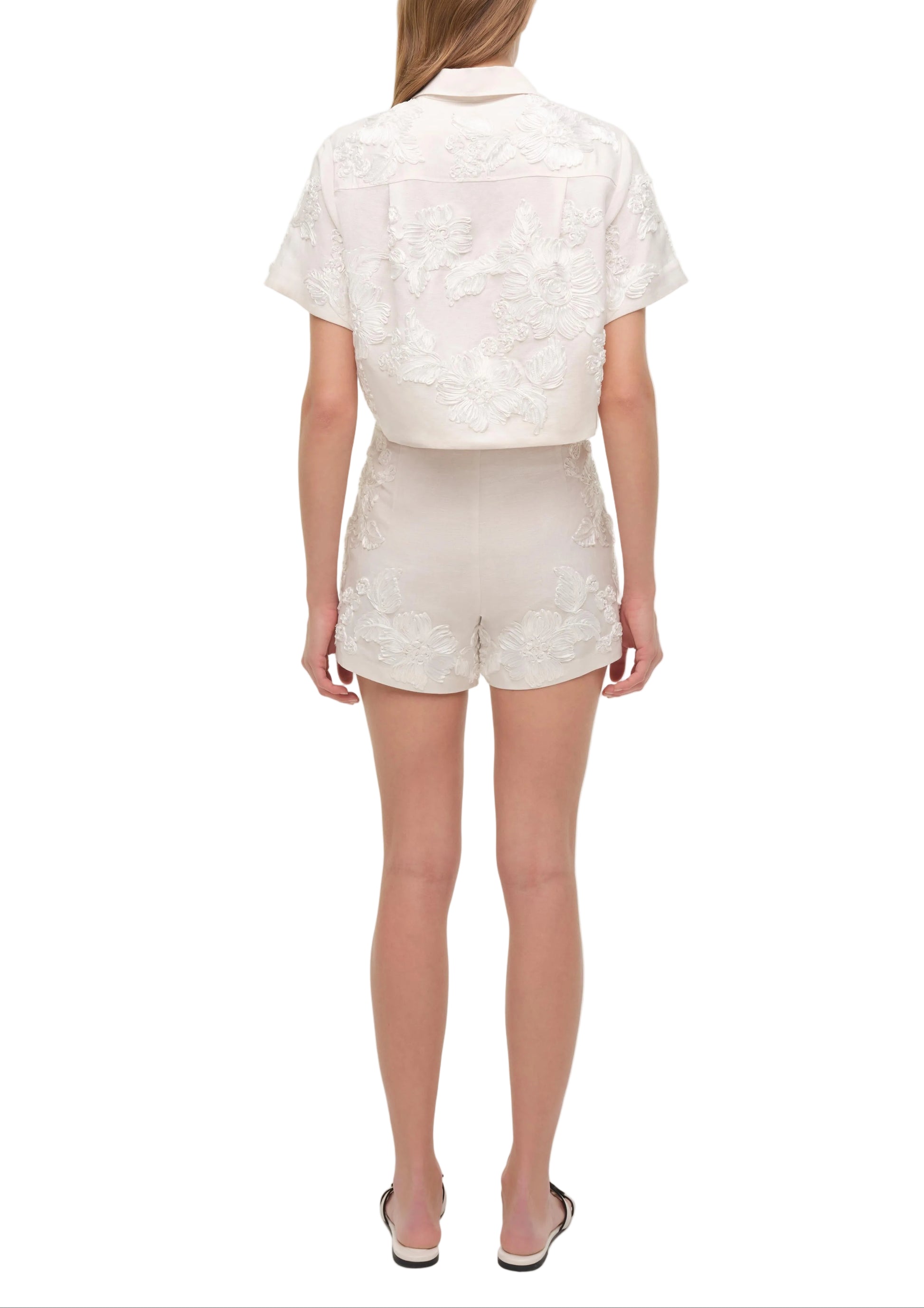 A woman faces away wearing the Simkhai Callie Camp Shirt and matching shorts with white slide sandals, creating a chic vacation look against a white background.