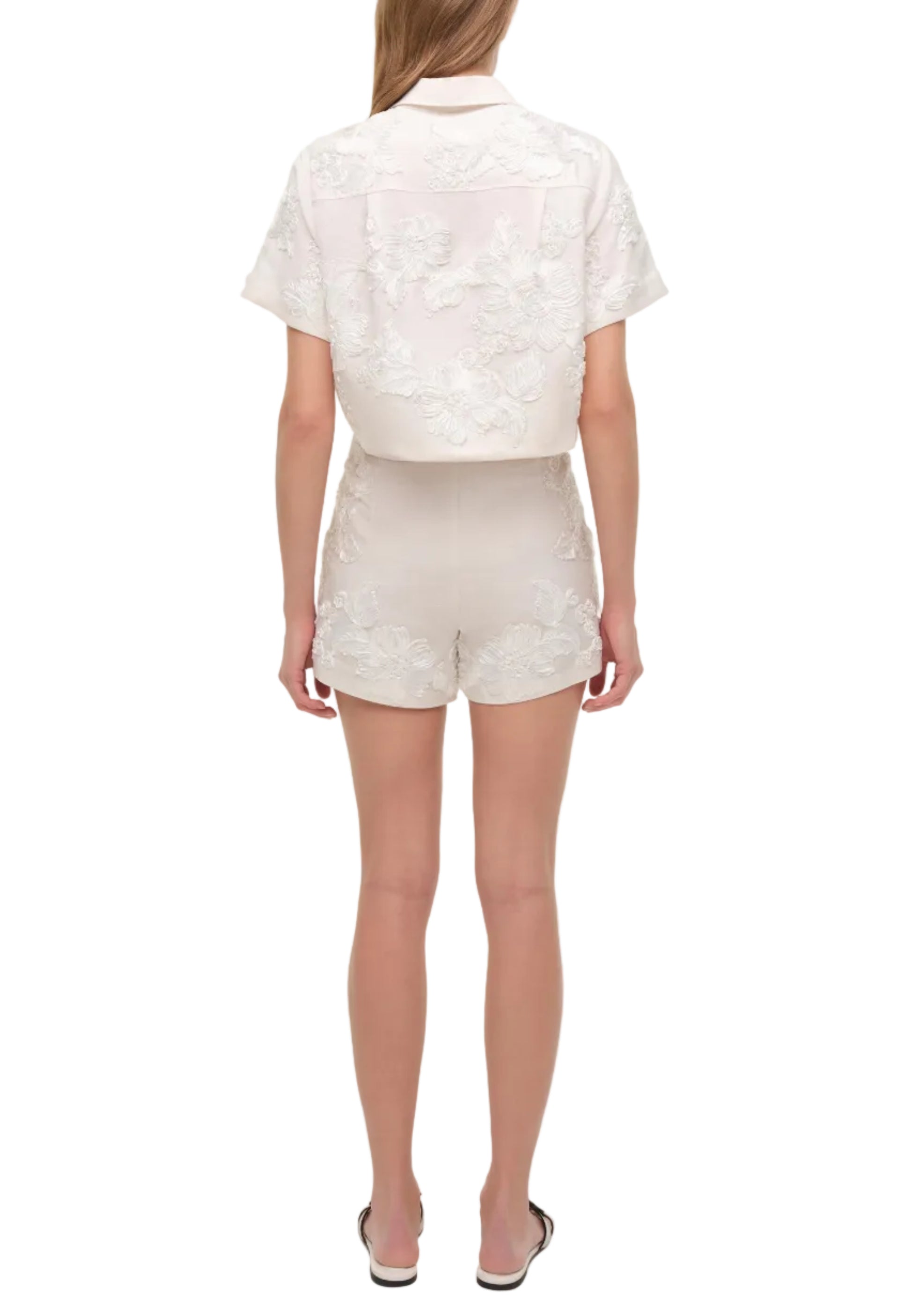 A woman stands facing away, wearing the Simkhai Sammie Short with a matching white embroidered short-sleeve shirt and white sandals.