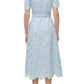 A woman faces away, wearing the Simkhai Audria Belted Midi Dress in light blue linen with white lace details and a matching belt, paired with white heeled sandals.