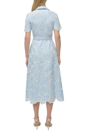 A woman faces away, wearing the Simkhai Audria Belted Midi Dress in light blue linen with white lace details and a matching belt, paired with white heeled sandals.