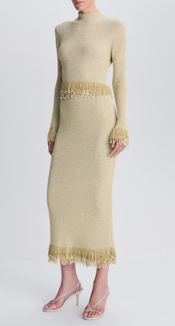 A Simkhai Zeni Midi Skirt, styled as a long-sleeved, high-neck metallic bouclé midi dress with pearl-trimmed fringe at the waist, cuffs, and hem, paired with thin-strapped white heels.