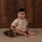 A baby wearing the Rylee & Cru Baby Relaxed Bubble Romper sits on a rug next to two large pine cones, with a wooden wall in the background—perfect for easy dressing and quick changes.