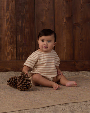 A baby wearing the Rylee & Cru Baby Relaxed Bubble Romper sits on a rug next to two large pine cones, with a wooden wall in the background—perfect for easy dressing and quick changes.