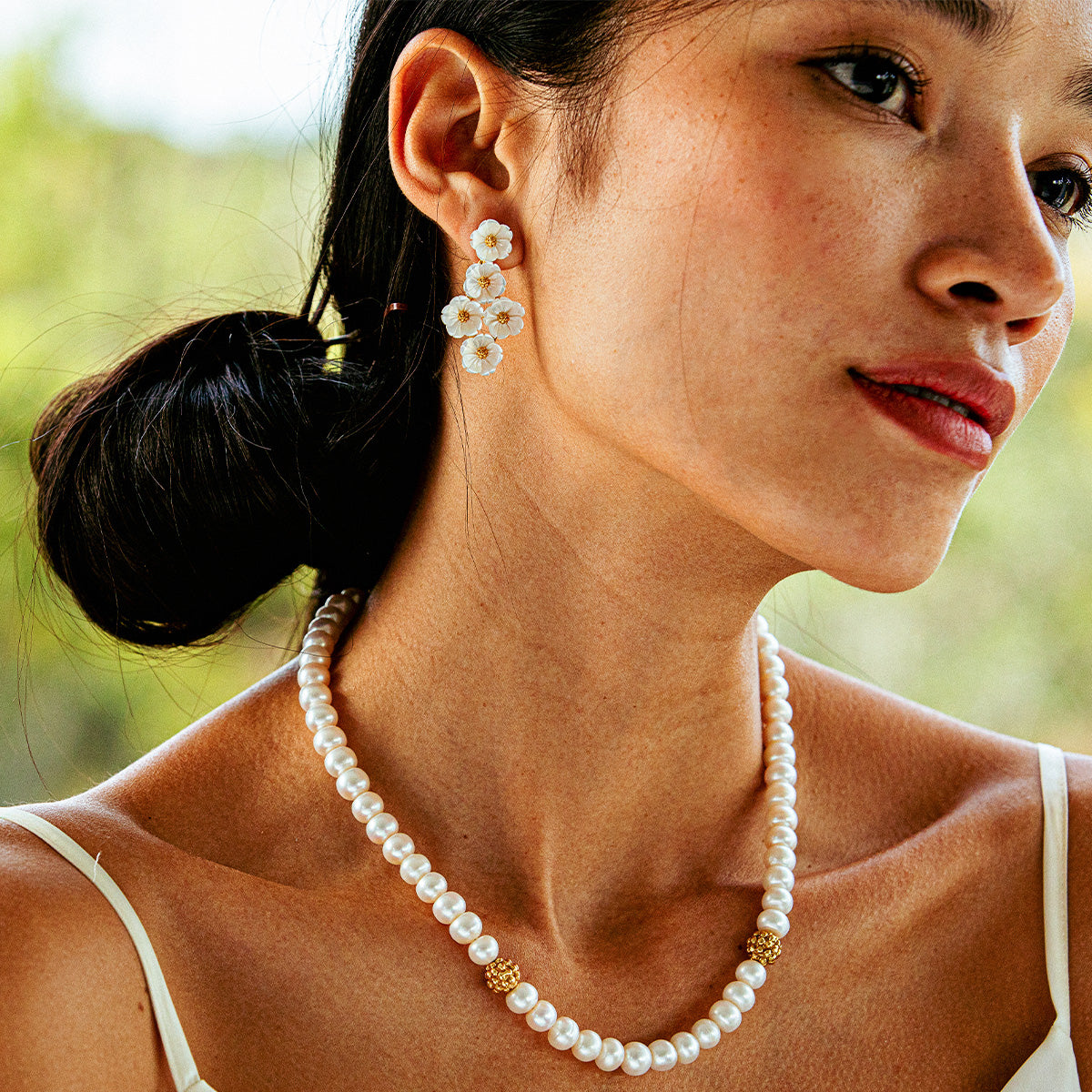 A woman wearing Juliska Capucine De Wulf's Mermaid Garden Floral Petite Post Drop Earrings and a pearl necklace with gold accents stands in a white tank top against a blurred green background.