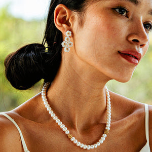 A woman wearing Juliska Capucine De Wulf's Mermaid Garden Floral Petite Post Drop Earrings and a pearl necklace with gold accents stands in a white tank top against a blurred green background.