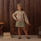 A young girl stands indoors on a rug, wearing a brown flower-patterned sweater with embroidered stitching, the Rylee & Cru Girls’ Denver Mini Skirt in green, and a headband, with one hand on her hip; wooden wall, books, bench, and lantern in the background.