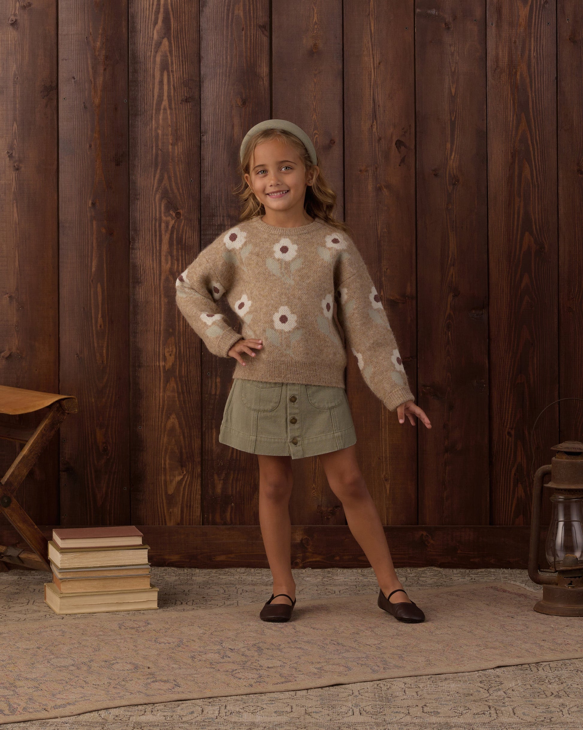 A young girl stands indoors on a rug, wearing a brown flower-patterned sweater with embroidered stitching, the Rylee & Cru Girls’ Denver Mini Skirt in green, and a headband, with one hand on her hip; wooden wall, books, bench, and lantern in the background.