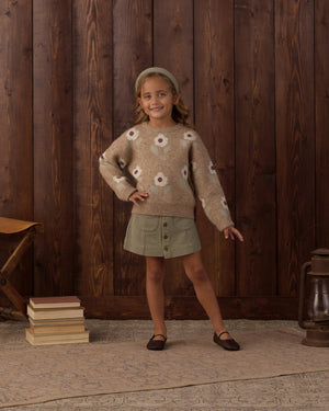 A young girl stands indoors on a rug, wearing a brown flower-patterned sweater with embroidered stitching, the Rylee & Cru Girls’ Denver Mini Skirt in green, and a headband, with one hand on her hip; wooden wall, books, bench, and lantern in the background.