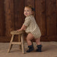 A baby wearing the Rylee & Cru Baby Girls’ Liv Romper, featuring a vintage-inspired design with a scalloped collar, stands in dark socks holding a small wooden stool, with a wooden wall and rug in the background.