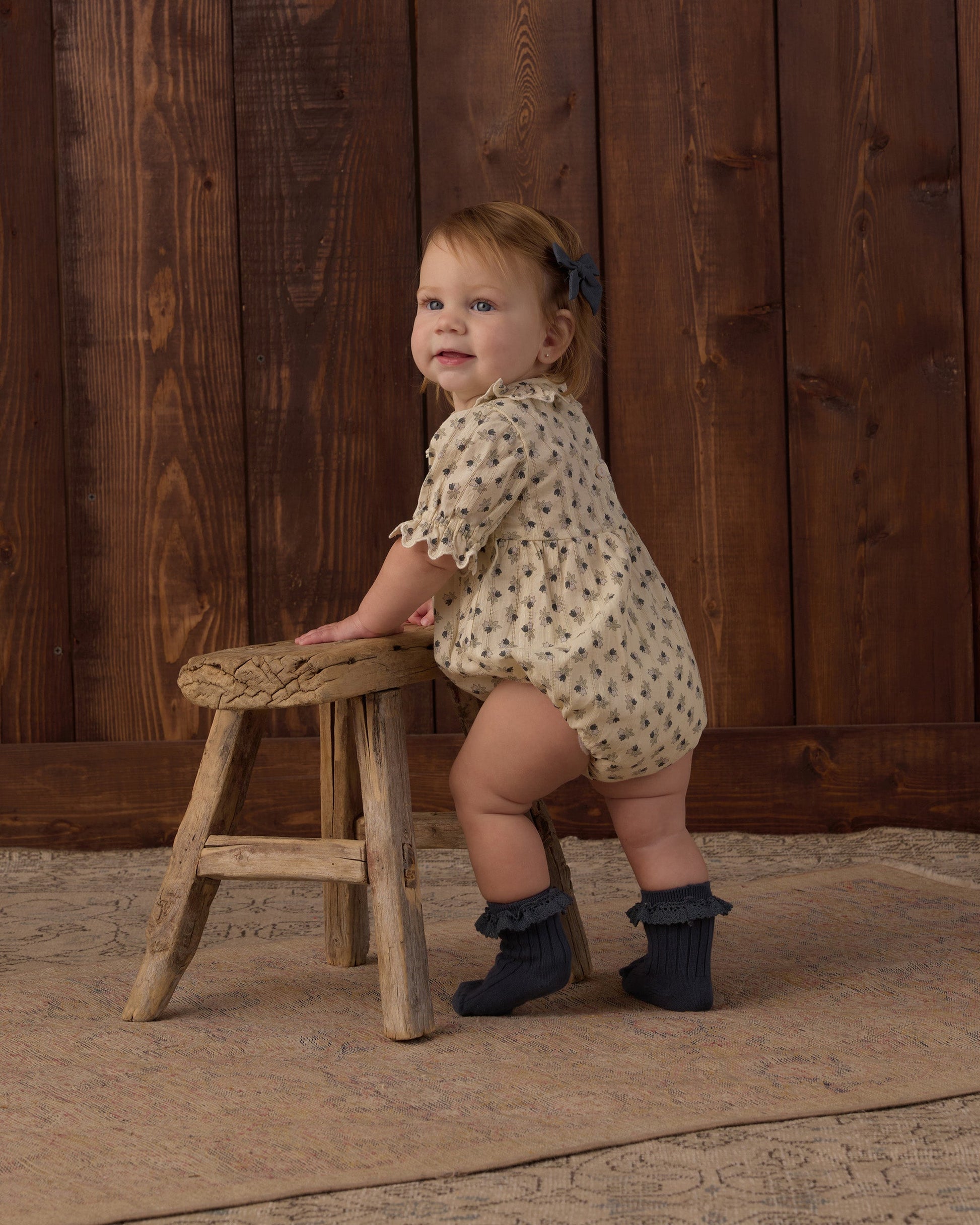 A baby wearing the Rylee & Cru Baby Girls’ Liv Romper, featuring a vintage-inspired design with a scalloped collar, stands in dark socks holding a small wooden stool, with a wooden wall and rug in the background.