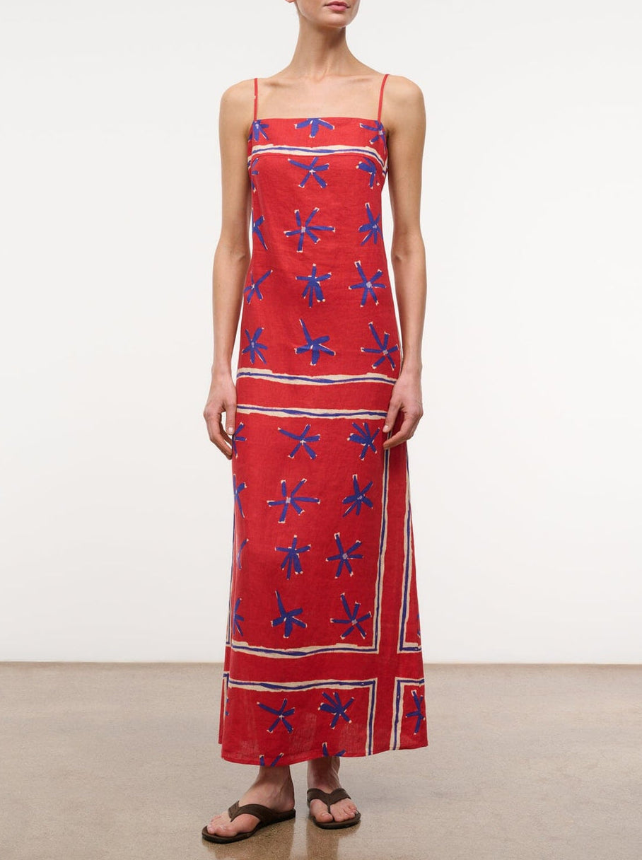 A person wears the Laura Linen Maxi Dress by Staud—a red sleeveless summer dress with blue star patterns and white stripes—paired with black flip-flops, standing against a plain background.