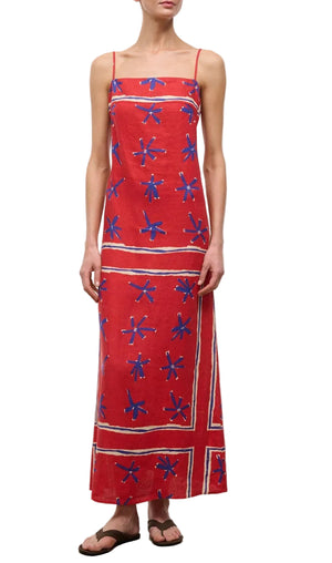 A person wears the Laura Linen Maxi Dress by Staud—a red sleeveless summer dress with blue star patterns and white stripes—paired with black flip-flops, standing against a plain background.