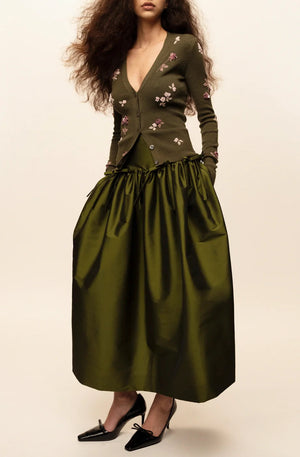 A person wears a dark lime Bernadette Oscar skirt, a matching embroidered cardigan, and black heeled pumps.