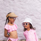 Two young girls in pink swimsuits and sunglasses stand before a light purple wall; one wears Snapper Rock's Lala Braid Visor for sun protection, while the other sports a white hat.