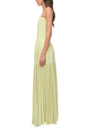 A woman stands in profile wearing the Simkhai Sandrine Dress, a strapless light yellow maxi with an elongated silhouette, fitted paneled bodice, and pleated skirt.